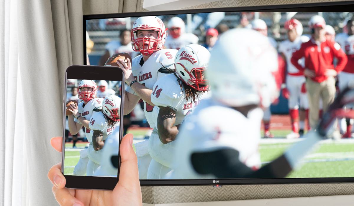 A hand holding an iPhone that is mirroring an image of a football match to an LG TV