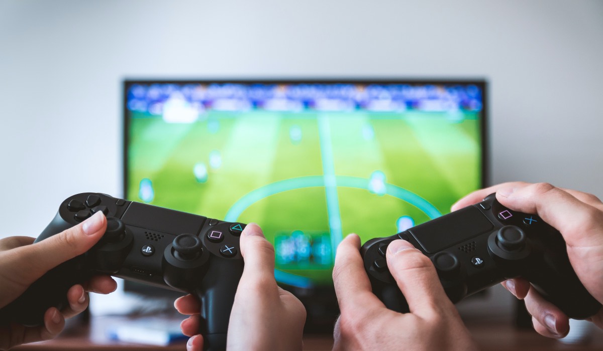 Two sets of hands holding a controller each. An LG TV with FIFA football video game on the screen